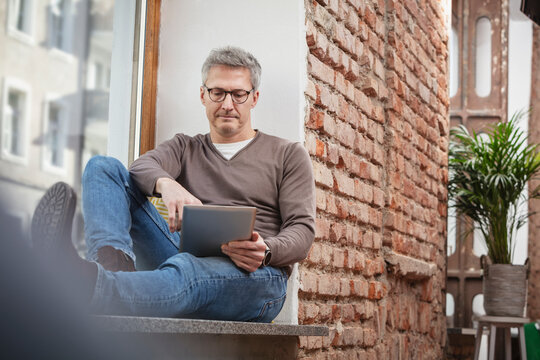 Mature man in eyeglasses using digital tablet while sitting on window sill at home