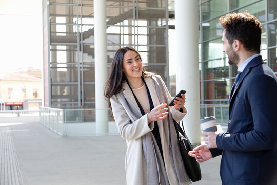 Smiling businesswoman with mobile phone talking to colleague on footpath