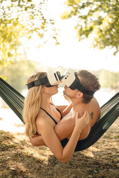 Couple relaxing in hammock by the lake, kissing, using VR goggles