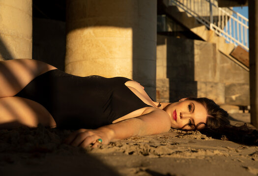 Smiling fashion model in one piece swimsuit lying on sand during sunset