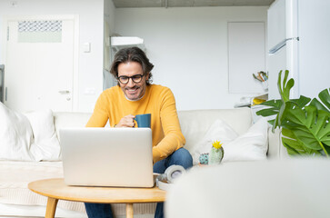 Smiling man holding mug while using laptop at home