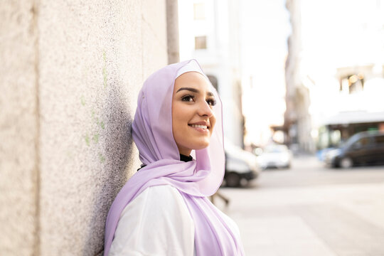Beautiful teenage girl in hijab looking away while standing by wall in city