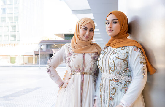 Beautiful friends in hijabs and dress standing together by wall