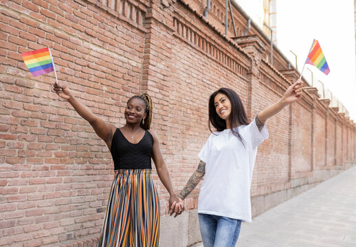 Multi-ethnic lesbian couple holding flag while standing on footpath near wall