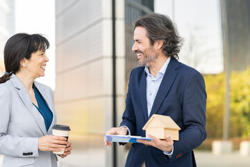 Smiling male real estate agent with house model discussing with female customer holding disposable cup in park