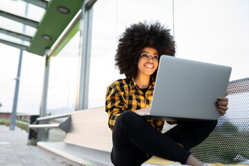 Smiling woman using laptop while sitting cross-legged on bench