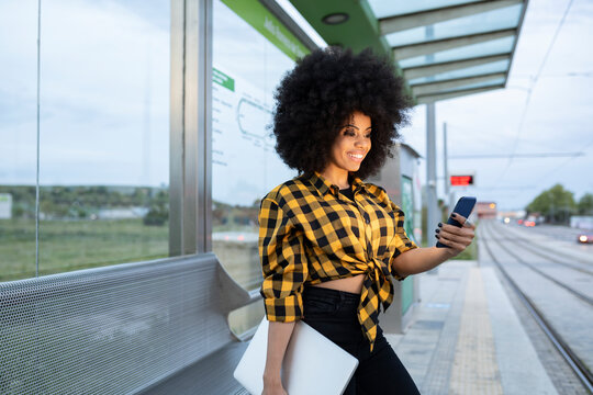 Smiling woman holding laptop while using smart phone at railroad station