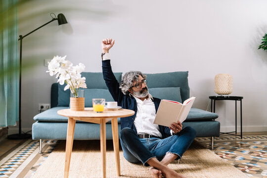Tired senior man stretching arm while reading book by sofa at home