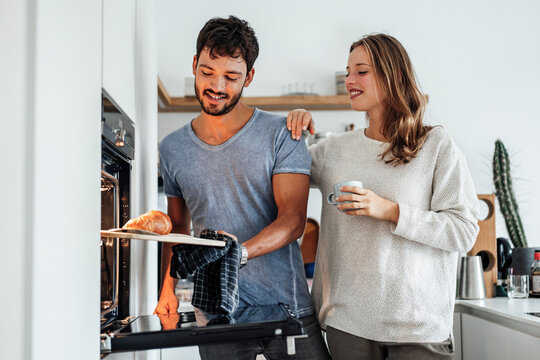 Smiling woman looking at boyfriend removing baked croissant from oven in kitchen