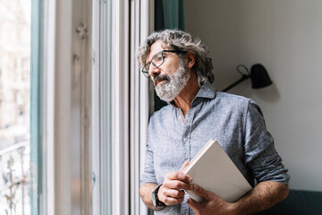 Thoughtful senior man holding book while looking through window at home