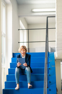 Smiling female owner using digital tablet while sitting on steps in workshop