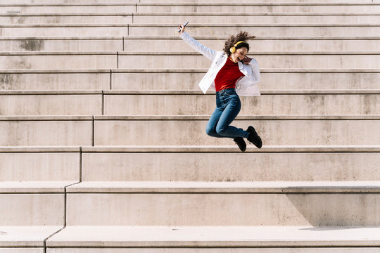 Ecstatic woman with headphones and smart phone jumping on staircase during sunny day