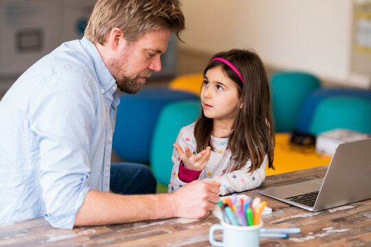 Girl gesturing while talking with father during e-learning at table