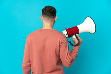 Young caucasian handsome man isolated on blue background holding a megaphone and in back position