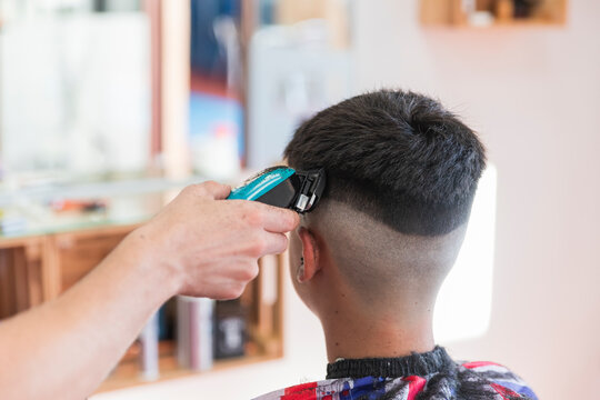 Male hairdresser using machine to do hairstyle of teenage boy at barber shop