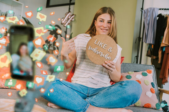 Female influencer showing like and subscribe cardboard while vlogging through mobile phone at home