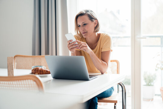 Smiling female professional with laptop using mobile phone at home