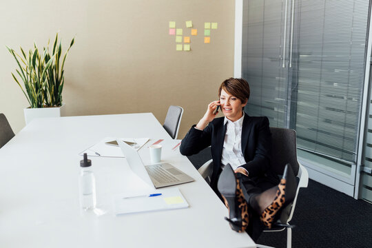 Smiling businesswoman looking at laptop while talking on mobile phone in office