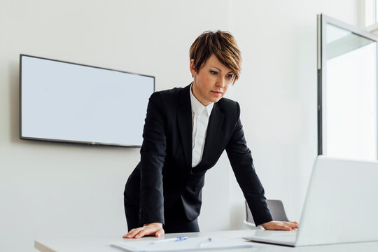 Thoughtful female business professional looking at laptop while leaning on desk in board room