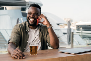 Young african man drinking beer at brewery bar using mobile phone with luxury yacht port in...