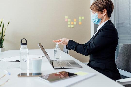 Female entrepreneur with protective face mask using hand sanitizer while sitting at desk
