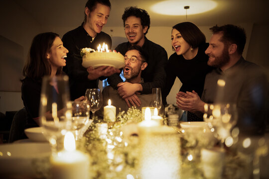 Surprised man looking at birthday cake held by male friend during celebration