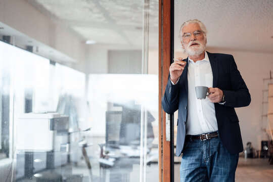 Senior businessman holding cigarette with coffee cup while leaning on glass wall at office