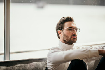 Relaxed man wearing eyeglasses sitting in front of bright window at home