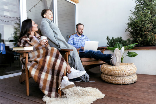 Mother and son wrapped in blanket while sitting with father using laptop on balcony