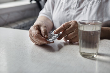 Sad old woman taking pills, health problems in old age, expensive medications. An elderly woman's hands unpacking several pills for taking medication. Grandma takes tablet and drinks a glass of water