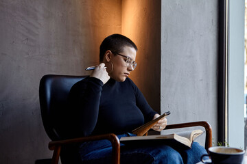 A young girl with glasses, with short hair is sitting in a cafe and reading a book. The girl looks...