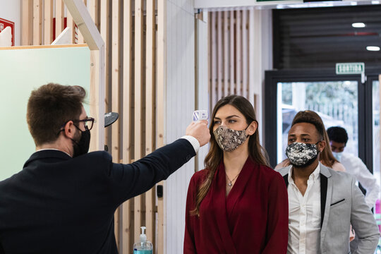 Man checking temperature of employees with face mask through infrared thermometer at office