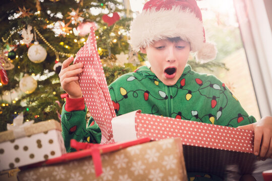 Excited boy in Santa hat unwrapping gift with Christmas tree in background at home