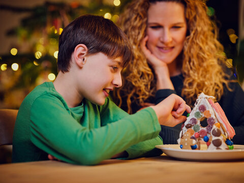 Smiling cute boy decorating gingerbread house by mother during Christmas