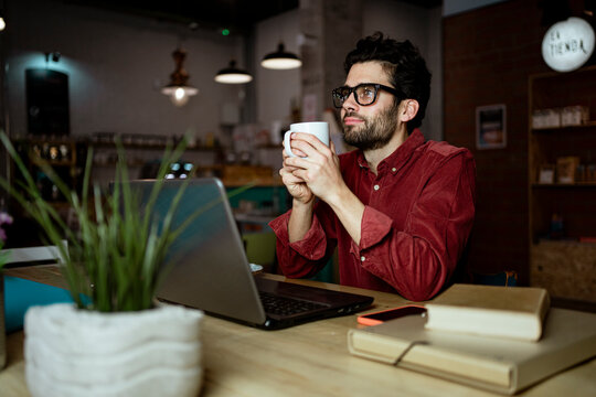 Thoughtful male freelance worker with laptop having coffee in illuminated cafe