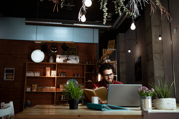 Male freelancer holding book while working on laptop in illuminated coffee shop