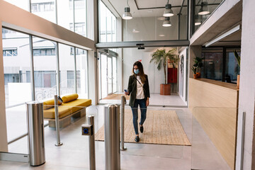 Businesswoman with protective face mask using ID card at turnstile while entering office