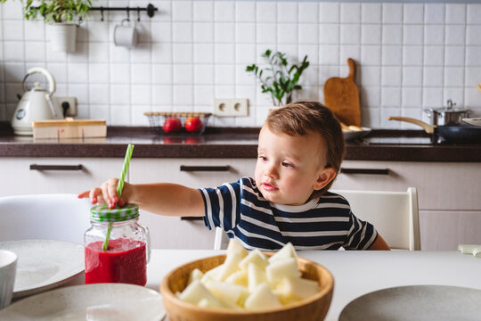 Cute boy holding straw in mason jar while sitting at dining table in kitchen
