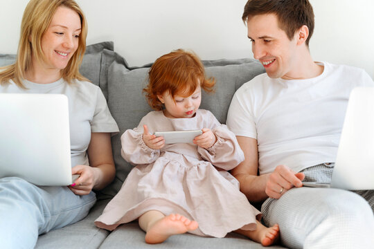 Smiling male and female entrepreneurs looking at daughter using mobile phone on sofa at home