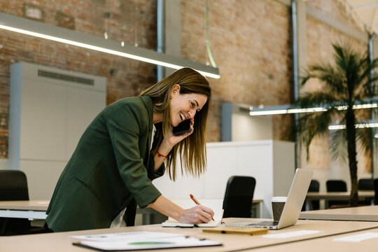 Cheerful businesswoman talking on smart phone while writing in diary by laptop at office