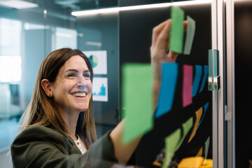 Excited businesswoman brainstorming using sticky notes on glass wall at office