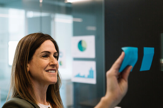 Smiling businesswoman sticking adhesive note on glass wall at office