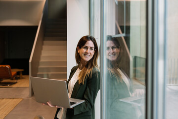 Thoughtful businesswoman holding laptop while looking through glass window at office