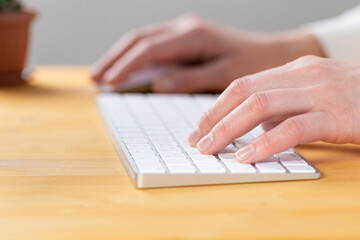An image of a woman's hand is typing on a white keyboard on a wooden table. Selective focus