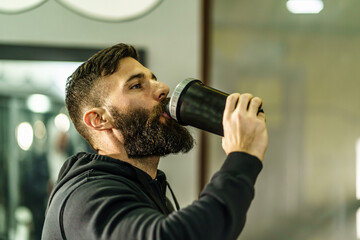 Young adult man with beard drinking bottle supplement shake in training at gym side view copy space...