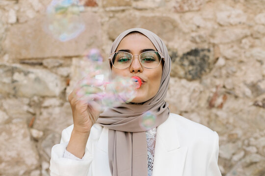 Young woman wearing headscarf blowing bubbles while standing in front of wall