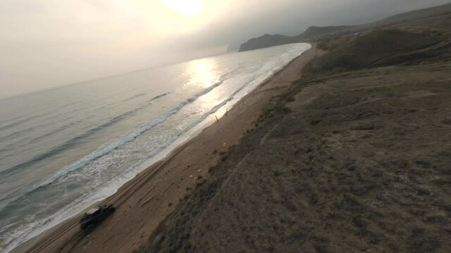 Large buggy vehicles drive along wet sand on coastline between brown hilly landscape and endless blue waving sea aerial fpv view