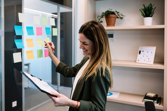 Smiling mid adult businesswoman holding clipboard while planning strategy on adhesive note at office