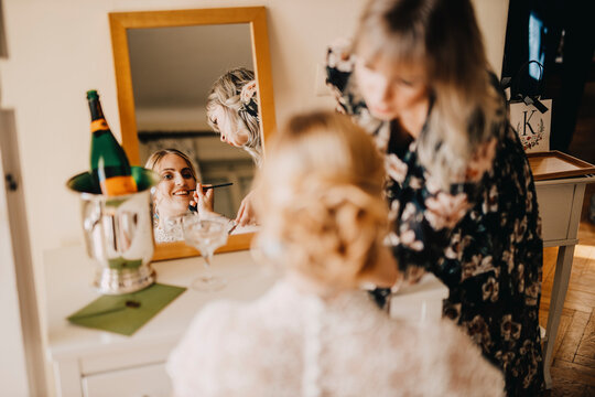 Bride looking in mirror while beautician applying make-up during wedding