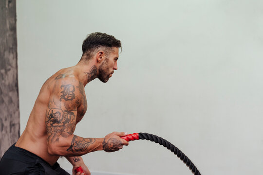 Shirtless sportsman training with rope by wall in gym